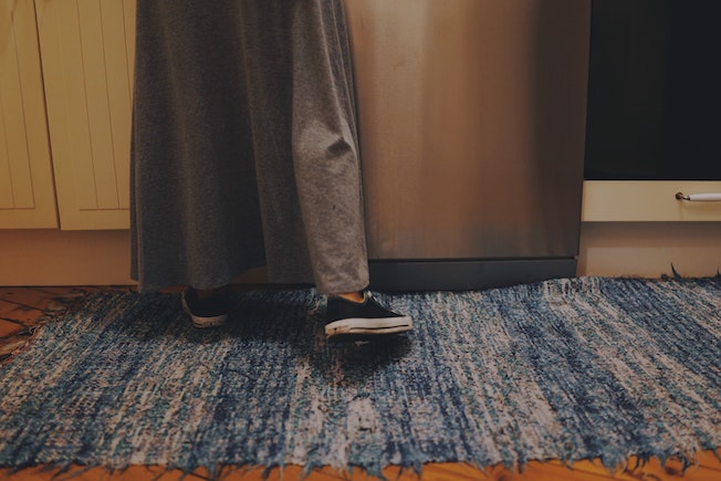 Holzfußboden mit einem blauen Teppich. Darauf steht eine Person, mit schwarzen Converse Schuhen und einem grauen langen Rock.