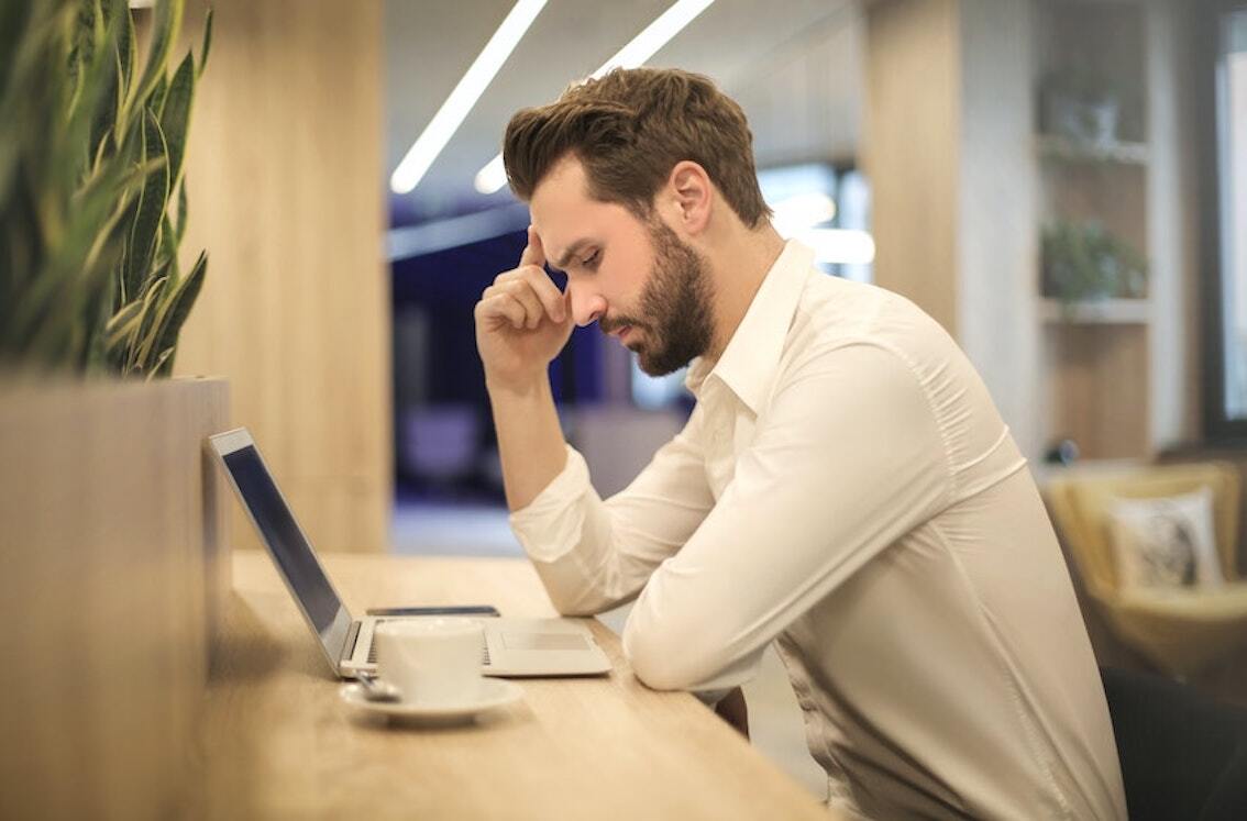 junger Mann sitzt nachdenklich an einem Tisch vor seinem Laptop.